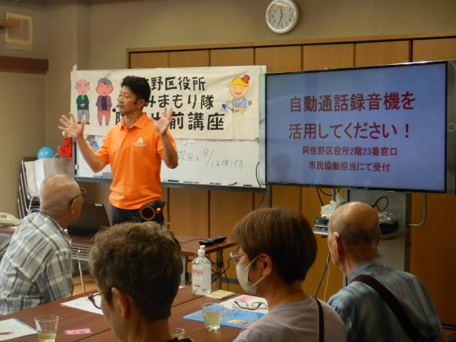 自動通話録音機を紹介し被害防止のため活用してくださいと呼びかける区役所職員
