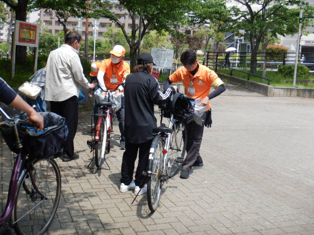 公園内でキャンペーン参加者の自転車前かごにひったくり防止カバー取りつける区役所職員