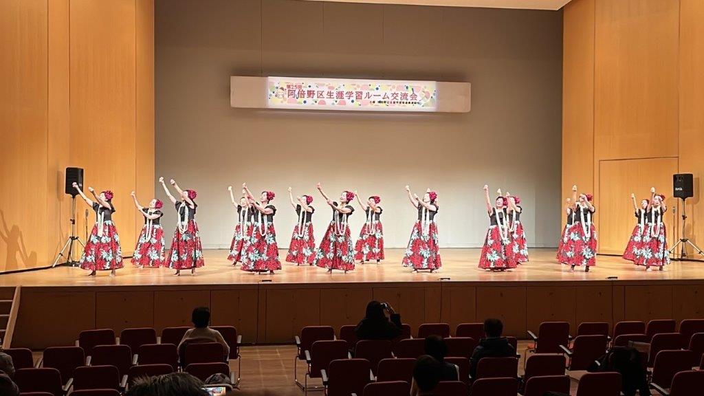 舞台上で衣装を着てレイをつけた16名がフラダンスをしている様子