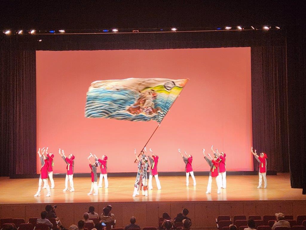 舞台上で法被を着た11名が鳴子を鳴らしながらよさこいを踊っている様子