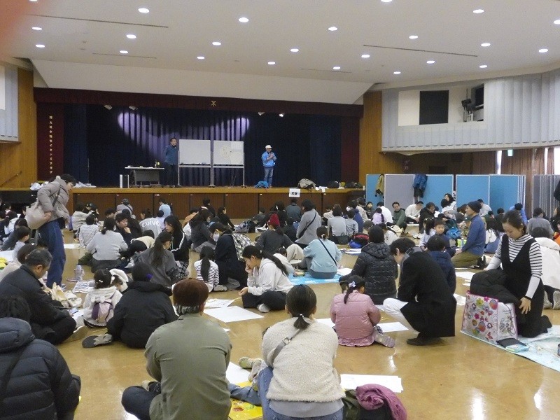 たくさんの人がたこつくりの会場ですわって説明を聞いている様子