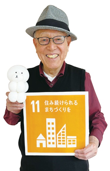代表の西方穂積さんがSDGsの11番のボードとバルーンアートで作った白い犬の作品を持っている写真