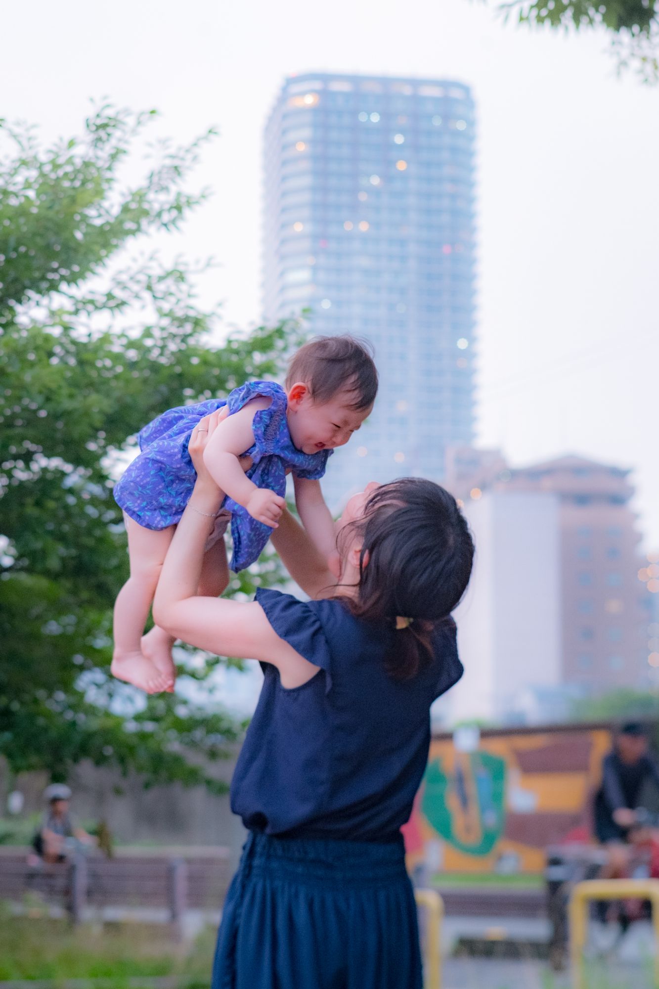 ビルの見える公園で女性が子どもを頭上に持ち上げている写真