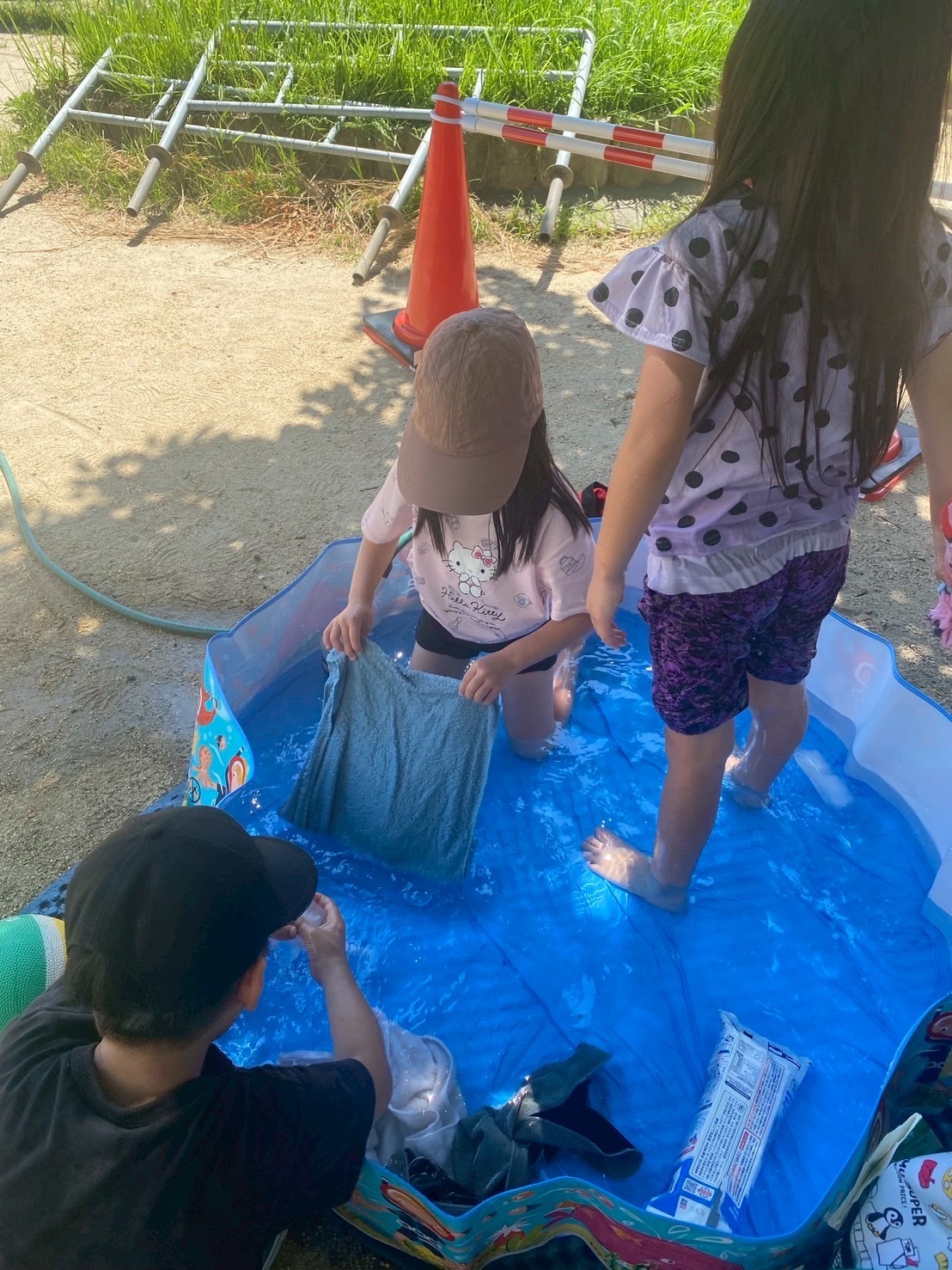 子どもたちが日陰で簡易プールに張った水で水遊びしている写真