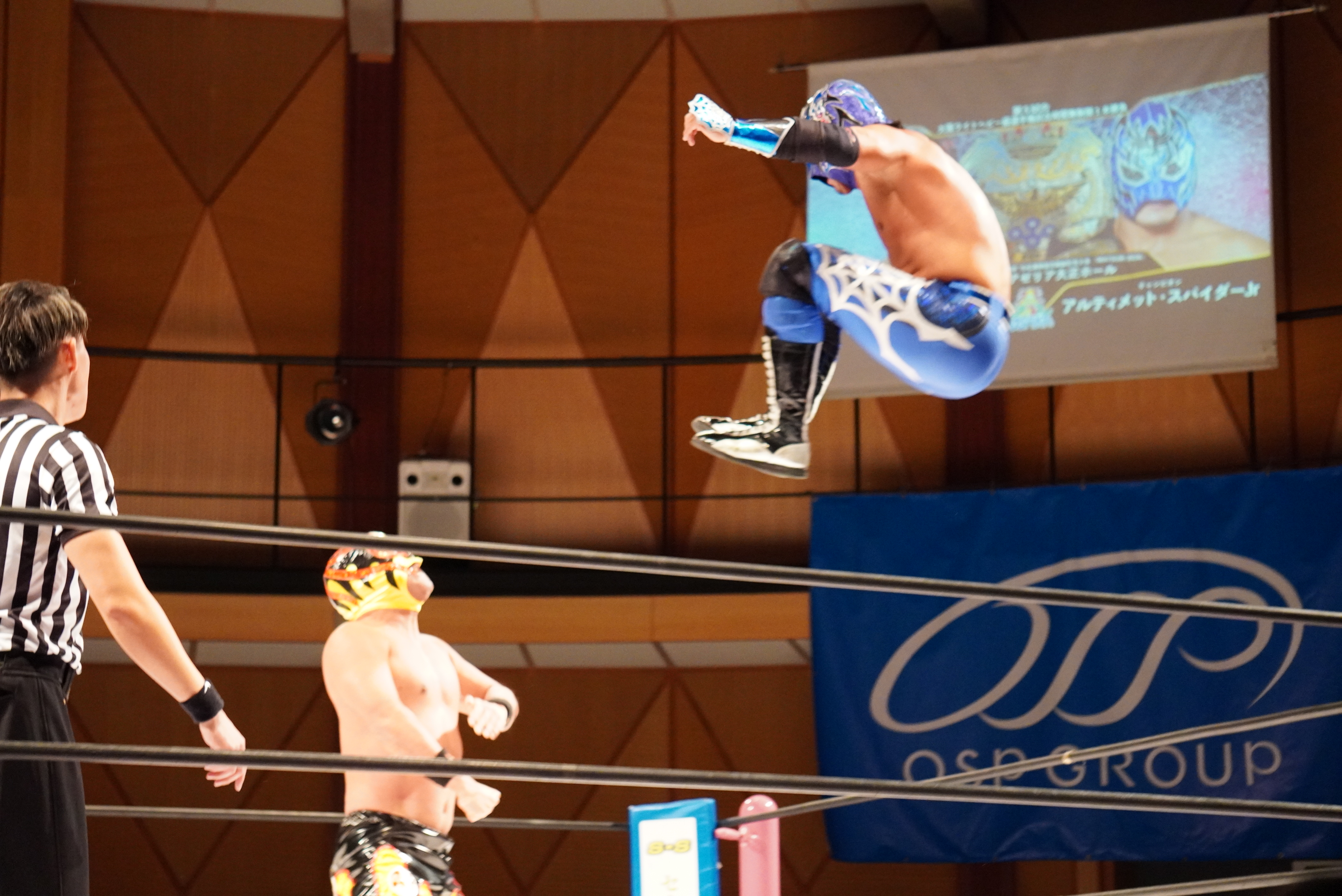 リング上で青いマスクのレスラーがジャンプして飛び技を放ち、下の黄色いマスクのレスラーが構える様子